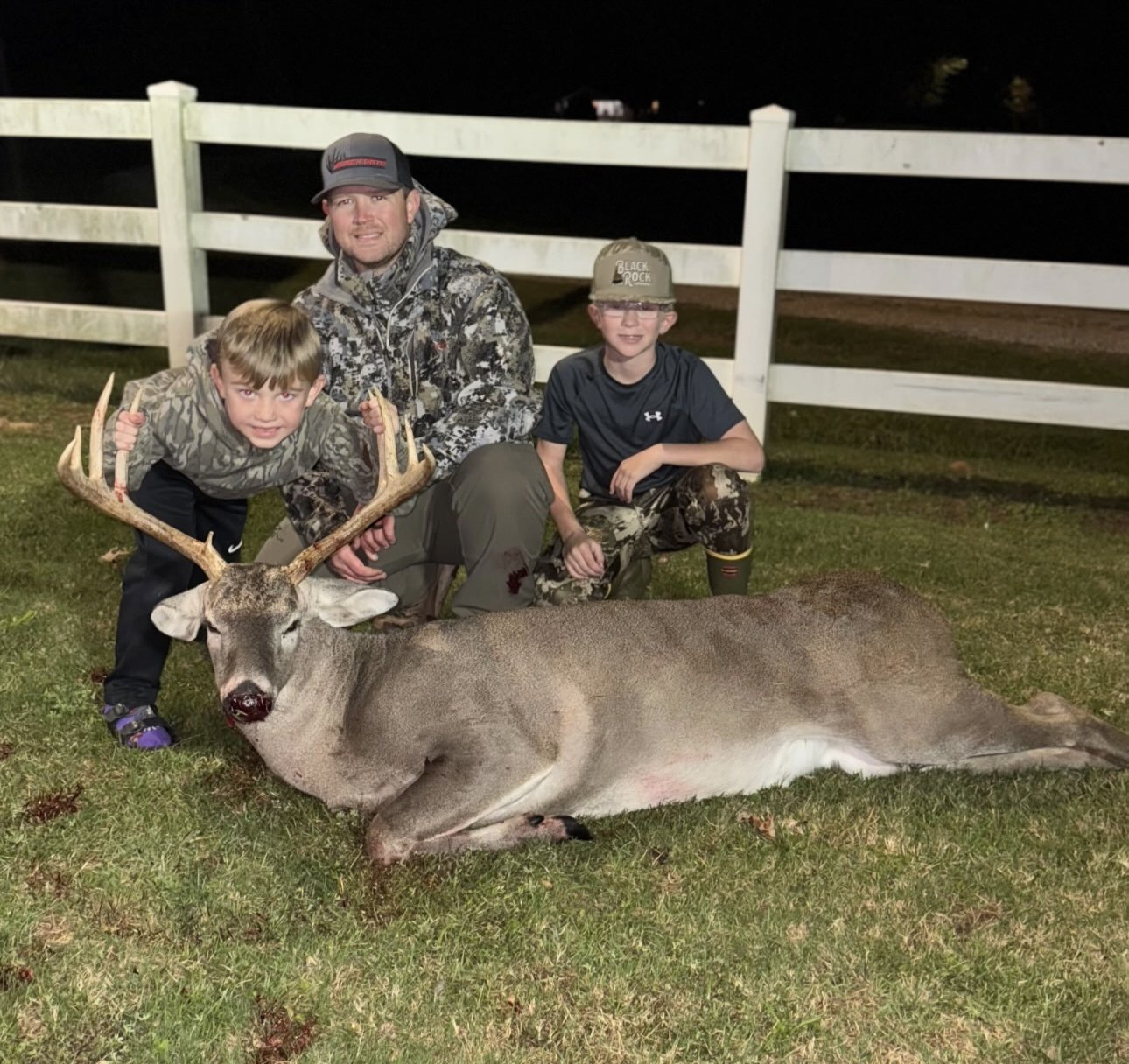 Two hunters with a buck beside a white fence line